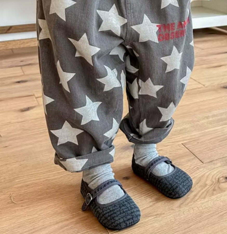 Child wearing gray pants with white stars and black shoes on a wooden floor.