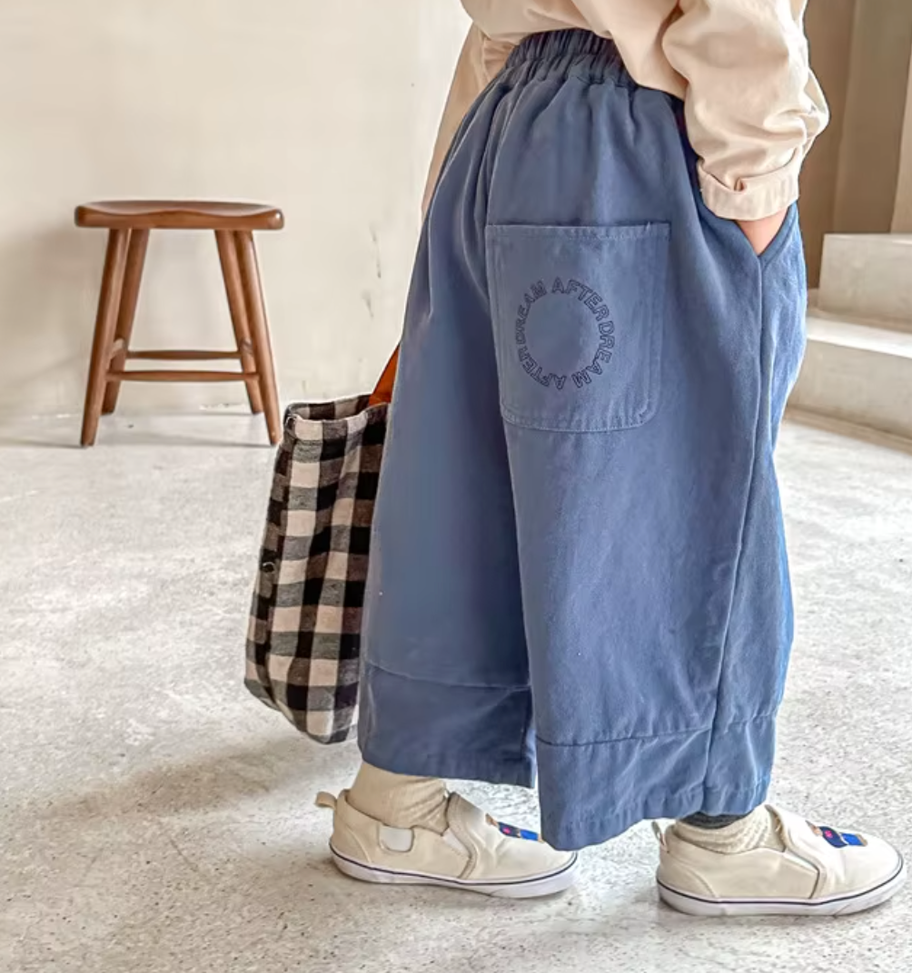 Person wearing blue pants with a visible brand logo, standing in a room with a wooden stool and checkered bag.